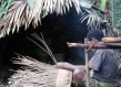 Man weaving basket beside a hut. 